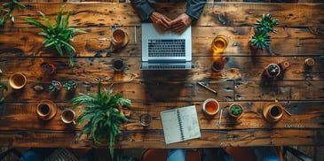 Top view of a wooden table with a laptop, notebooks, coffee cups, and various potted plants.